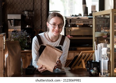 Young Smiling Woman (salesperson In Bakery) Holding Craft Paper Packing Bag For Taking Away Food In Coffee Shop