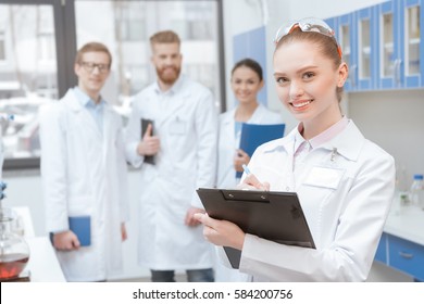 Young Smiling Woman Scientist With Clipboard Standing Near Colleagues In Lab