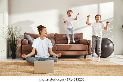 Young Smiling Woman Sitting In Lotus Pose With Laptop On Floor And Calming Down While Noisy Kids Jumping Around In Living Room
