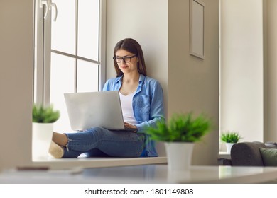 Young smiling woman sitting on windows and working online on laptop - Shutterstock ID 1801149808