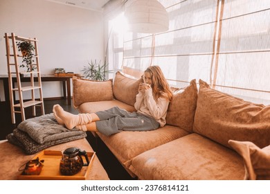 Young smiling woman sitting on sofa and looking up while drinking hot tea. Young millennial woman thinking at home in leisure time. Happy girl relaxing at home on bright winter morning - Powered by Shutterstock