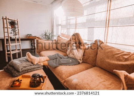 Young smiling woman sitting on sofa and looking up while drinking hot tea. Young millennial woman thinking at home in leisure time. Happy girl relaxing at home on bright winter morning