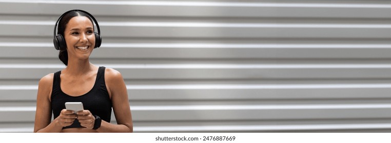 Young Smiling Woman In Sportswear And Wireless Headphones Resting With Smartphone After Training Outdoors, Fit Lady Standing Near Urban Wall, Enjoying Music Playlist For Running, Copy Space - Powered by Shutterstock