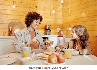 Young smiling woman spreading peanut butter on slice of wheat bread while sitting by served table and making sandwich for her son - Powered by Shutterstock