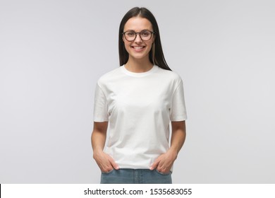 Young smiling woman standing with hands in pockets, wearing blank white tshirt with copy space, isolated on gray background