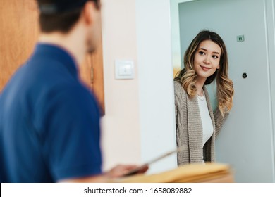 Young Smiling Woman Standing On A Doorway While Receiving Home Delivery From A Courier. 