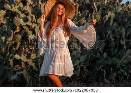 Young smiling  woman in straw hat and white dress  looking at camera standing on background of exotic cactuses