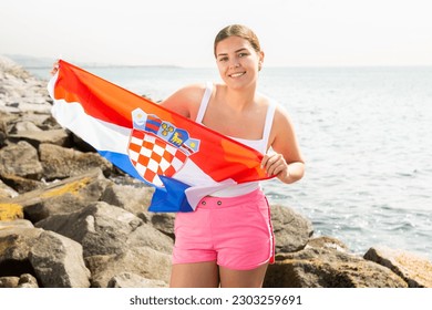 Young smiling woman in summer clothes posing with big flag of Croatia on sea coast
