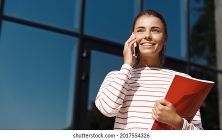 Young Smiling Woman Talking On Mobile Phone, Standing Outside Building With Book In Her Hand