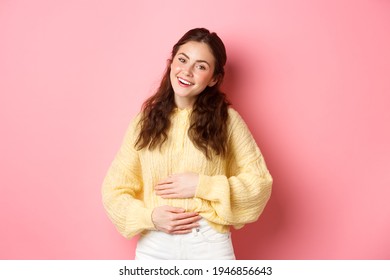 Young smiling woman touching her stomach with relieved, happy face, feeling good after eating yoghurt or medicine from painful cramps, standing against pink background