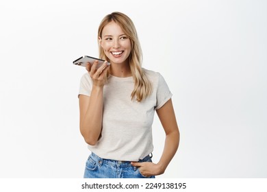 Young smiling woman using mobile phone speakerphone, translating her voice in smartphone app, recording herself, standing over white background - Powered by Shutterstock