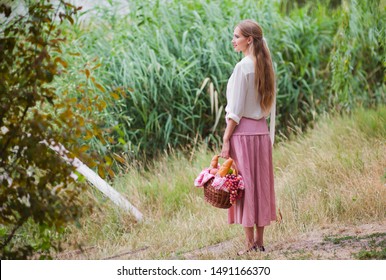 Young smiling woman in vintage retro style clothes holds a picnic basket in her hand against the background of reeds on the lake