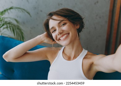 Young smiling woman wear white tank shirt do selfie shot pov on mobile cell phone sit on blue sofa stay at home hotel flat rest relax spend free spare time in living room indoor People lounge concept - Powered by Shutterstock