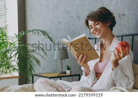 Young smiling woman wear white shirt pajama she lying in bed read book study eat apple rest relax spend time in bedroom lounge home in own room hotel wake up dream be lost in reverie good mood day.