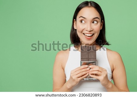 Young smiling woman wear white clothes hold in hand dark chocolate bar look aside on area isolated on plain pastel light green background. Proper nutrition healthy fast food unhealthy choice concept