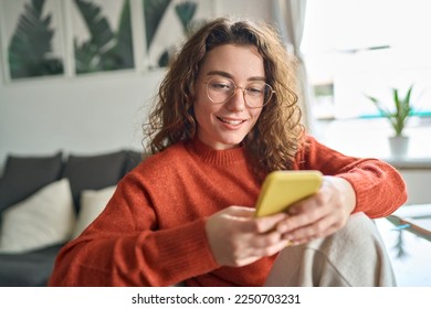Young smiling woman wearing glasses holding smartphone using cellphone modern technology, looking at mobile, checking cell phone apps, texting, browsing internet for shopping sitting at home.
