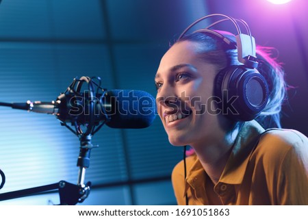 Young smiling woman wearing headphones and talking into a microphone at the radio station, entertainment and communication concept