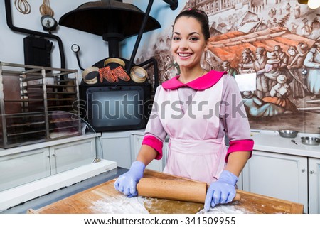 Young smiling woman, wearing in pink dress with ribbon in her hair, apron and violet gloves, kneads the dough on wooden board with rolling pin, in the kitchen with painted walls, waist up
