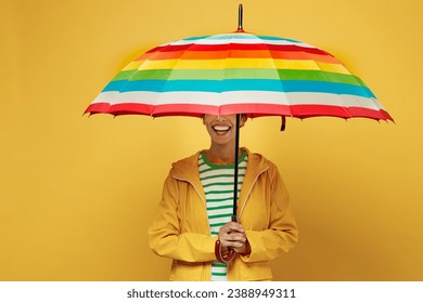 Young smiling woman wearing waterproof raincoat outerwear hold in hand cover face with striped umbrella isolated on plain yellow background studio. Outdoors lifestyle wet fall weather season concept - Powered by Shutterstock