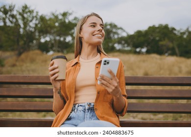 Young smiling woman wears orange shirt casual clothes use mobile cell phone drink coffee look aside sit on bench walk rest relax in spring green city park outdoors on nature. Urban lifestyle concept - Powered by Shutterstock