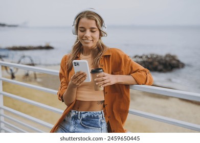 Young smiling woman wears orange shirt casual clothes headphones listen to music use mobile cell phone drink coffee walk on sea ocean sand shore beach outdoor seaside in summer day. Lifestyle concept - Powered by Shutterstock