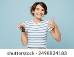 Young smiling woman wears striped t-shirt casual clothes hold in hand car key fob keyless system show thumb up isolated on plain pastel light blue cyan background studio portrait. Lifestyle concept