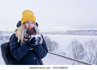 Young Smiling Woman In Yellow Beanie And Black Padded Jacket With Black Backpack Is Holding Snow In Her Hands Against The Winter Landscape With Icy River