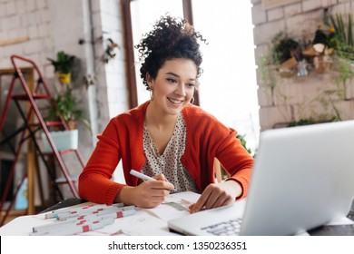 Jovem mulher sorridente com cabelo escuro encaracolado sentado à mesa feliz trabalhando no laptop desenhando ilustrações de moda passando o tempo na oficina moderna e aconchegante com grandes janelas