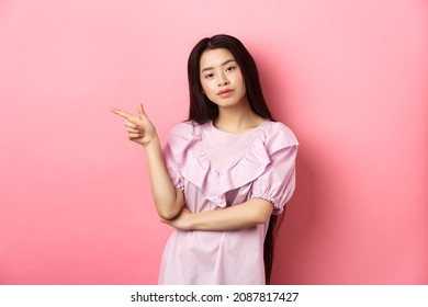 Young Smug Asian Girl Looking Cool And Pointing Finger Left At Logo, Advertising Product On Pink Romantic Background