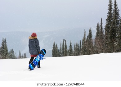 Young Snowboarder Girl Stand On Top Of A Mountain Turning Back To Camera Holding Her Snowboard And Watching Downhill Of A Mountain. Prepearing To Ride And Searching For Her Line. Freeride In Blizzard.