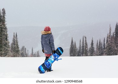 Young Snowboarder Girl Stand On Top Of A Mountain Turning Back To Camera Holding Her Snowboard And Watching Downhill Of A Mountain. Prepearing To Ride And Searching For Her Line. Freeride In Blizzard.