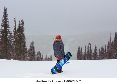 Young Snowboarder Girl Stand On Top Of A Mountain Turning Back To Camera Holding Her Snowboard And Watching Downhill Of A Mountain. Prepearing To Ride And Searching For Her Line. Freeride In Blizzard.