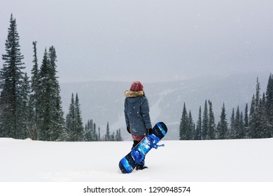 Young Snowboarder Girl Stand On Top Of A Mountain Turning Back To Camera Holding Her Snowboard And Watching Downhill Of A Mountain. Prepearing To Ride And Searching For Her Line. Freeride In Blizzard.