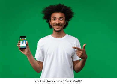 Young soccer fan man smiling and pointing with his index finger at smartphone with bookmaker's mobile application interface on its screen in hand. His bet played. Isolated over green background.