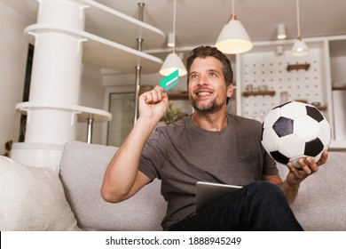 Young Soccer Fan Man Watching Football Game On Television At Living Room Couch. He Holds A Credit Card And A Digital Tablet For Online Betting.