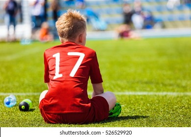 Young Soccer Football Player. Youth Athlete In Red Football Sportswear. Boy Sitting Alone On Soccer Pitch And Watching Youth Soccer Tournament. Youth Football Cup For Kids