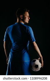 Young Soccer Player With Ball On Black Background In Studio. Football Player With A Stylish Haircut. Back View. Blue Shirt.