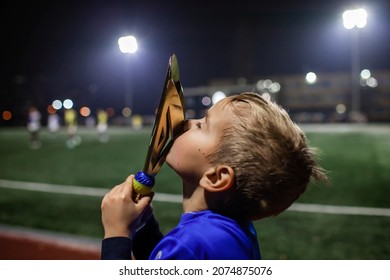 Young soccer player in blue jersey with ten number kissing a winners cup after the winning goal in the football tournament, illuminated stadium, sport, winner and success. Dreams come true