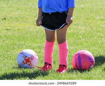 A young soccer player stands confidently on a grassy field, wearing a blue jersey, black shorts, and bright pink socks. Two colorful soccer balls rest on either side of her. - Powered by Shutterstock