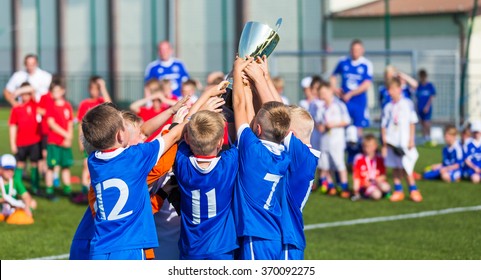 Young Soccer Players Holding Trophy. Boys Celebrating Soccer Football Championship. Winning Team Of Sport Tournament For Kids Children. Horizontal Sport Background.