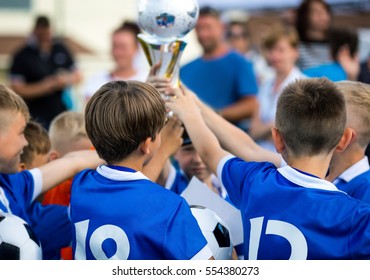 Young Soccer Players Holding Trophy. Children Soccer Football Champions. Boys Celebrating Soccer Championship. Winning Team Of Sport Kids Tournament. Youth Soccer Cup Winners Parents In The Background