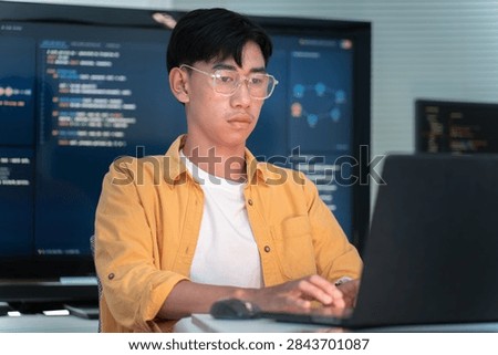 Young software developer wearing glasses codes on a laptop beside programming displays in a modern technology workspace