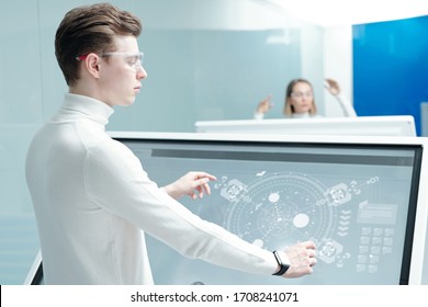 Young Software Engineer In Smart Goggles Standing By Large Interactive Display With Holographic Schemes While Analyzing Virtual Data