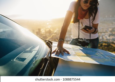 Young Solo Traveler Looking At Mobile Phone And Tourist Map On Car Hood. Young Female Tourist On A Road Trip Searching For Directions.