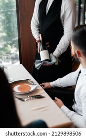 A Young Sommelier In A Stylish Apron Demonstrates And Offers Fine Wine To A Beautiful Couple In A Restaurant. Customer Service In An Elite Restaurant And A Public Restaurant.