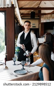 A Young Sommelier In A Stylish Apron Demonstrates And Offers Fine Wine To A Beautiful Couple In A Restaurant. Customer Service In An Elite Restaurant And A Public Restaurant.