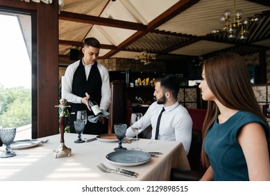 A Young Sommelier In A Stylish Apron Demonstrates And Offers Fine Wine To A Beautiful Couple In A Restaurant. Customer Service In An Elite Restaurant And A Public Restaurant.