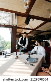 A Young Sommelier In A Stylish Apron Demonstrates And Offers Fine Wine To A Beautiful Couple In A Restaurant. Customer Service In An Elite Restaurant And A Public Restaurant.