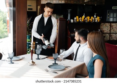 A Young Sommelier In A Stylish Apron Demonstrates And Offers Fine Wine To A Beautiful Couple In A Restaurant. Customer Service In An Elite Restaurant And A Public Restaurant.