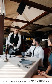 A Young Sommelier In A Stylish Apron Demonstrates And Offers Fine Wine To A Beautiful Couple In A Restaurant. Customer Service In An Elite Restaurant And A Public Restaurant.